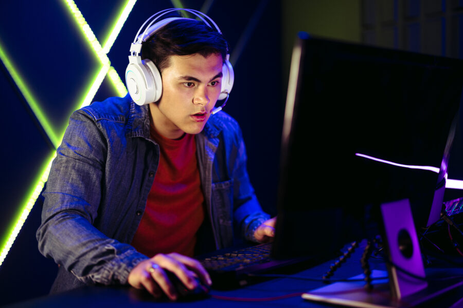 Young man focusing on the computer screen as he plays an online video game. With his ryes fixed on the moving images and hand gripping the mouse, he strategically navigates through the virtual world of the game.